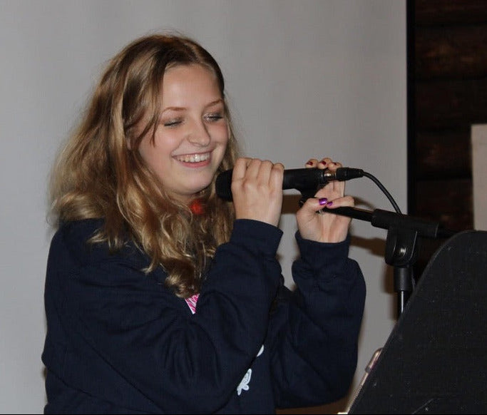 Reeceabella holding a microphone in front of a white screen. She is 14 here.
