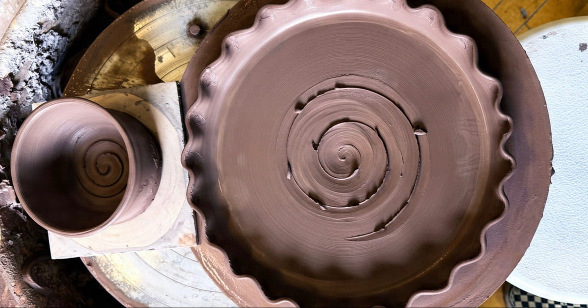 A top-down view of a pottery wheel with a large spiral-shaped clay vessel in motion and a smaller matching bowl to the left, set on a pottery wheel.