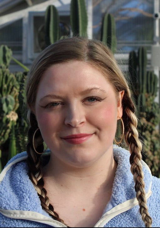 A young woman with blonde braids is wearing a light blue fleece zip-up hoodie with white trim, standing in front of a greenhouse filled with tall cacti.