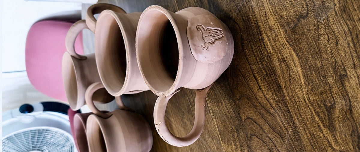 A group of handmade brown clay mugs and bowls arranged on a dark wooden table, with a white fan and pink chair visible in the background.