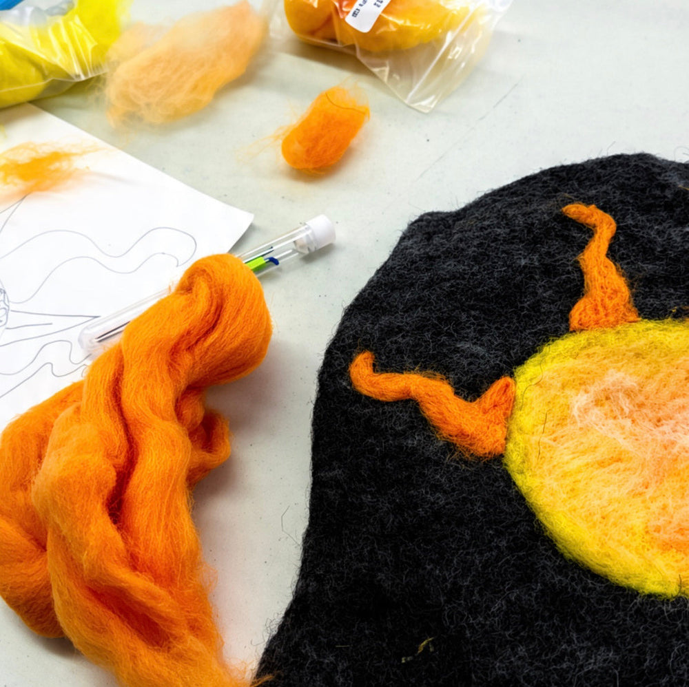 A close-up of a handcrafted felted wool sun on a white table, surrounded by orange and yellow wool fibers, a needle Felting tool, and a felted wool sun patch.
